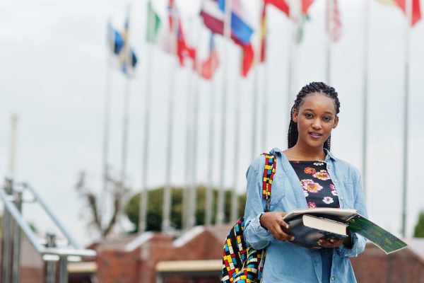 African student female posed with backpack and school items on yard of university, against flags of different countries.